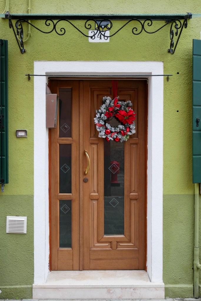In Burano a home is painted two shades of green - olive and avacado. The front door is wooden with a white trim and marble flooring. There's a green, white and red Christmas wreath on the front door.