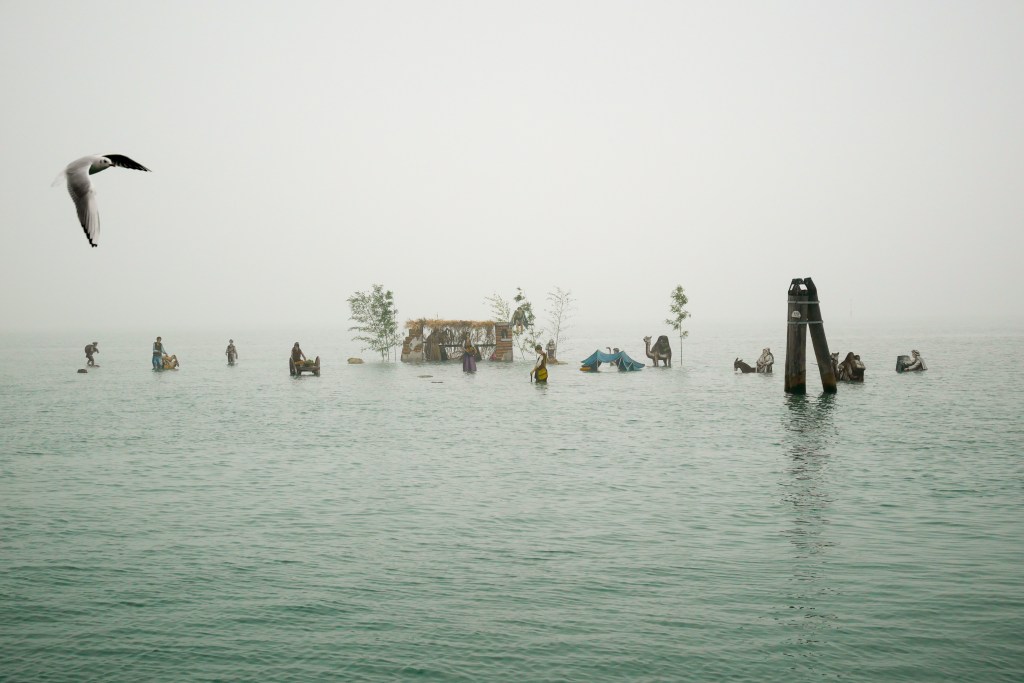 View of the Nativity on the Water in the Venetian Lagoon as seen from Burano. Plywood silhouettes are anchored in the sea, and depict shepherds, donkeys, a camel, etc. as well as an inn. A seagull flies overhead and is captured mid-flight. The sky is foggy so the horizon line is faded - we can't exactly tell where the greyish-blue sea stops or the grey sky begins.