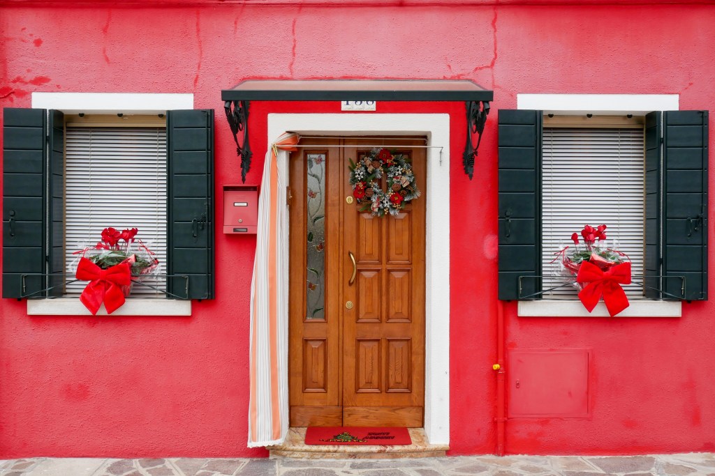 In Burano a house front is painted bright red. In the centre is a wooden door with white trim, and on either side is a window each with white trim, two opened black shutters, white blinds and a basket of red flowers with a large red bow sitting on the outside window sill.