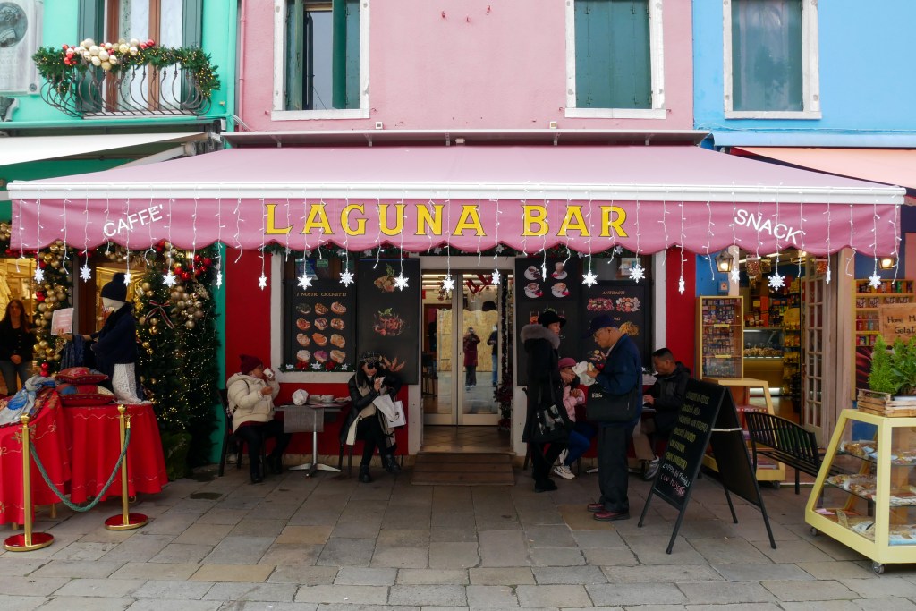 In Burano there's a restaurant with a red awning that says "LAGUNA BAR" in yellow letters.From the front of the awning hang icicle style Christmas lights with snowflakes at the ends, but it's daytime so the lights are not lit. Underneath the awning are two tables, one either side of the door and each with people sitting on chairs and eating, drinking and talking with each other. There's also an A-board sign with something written, but we can't make out what it says. Next to the board is a man waiting by himself. It's winter and everyone is bundled up in hats, scarves, and bulky winter coats.