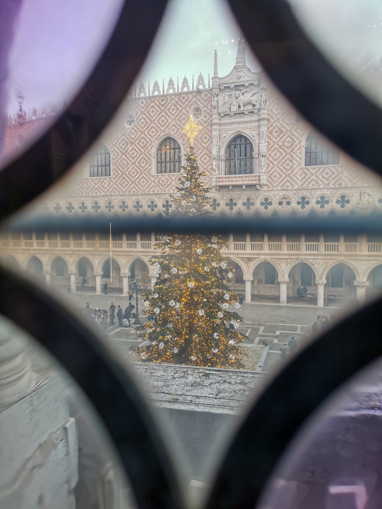 Inside a building on Piazza San Marco, looking through the decorative glass to the Christmas tree in front of Doge's Palace. The glass decorations create a diamond frame around the Christmas tree from the angle we're viewing.