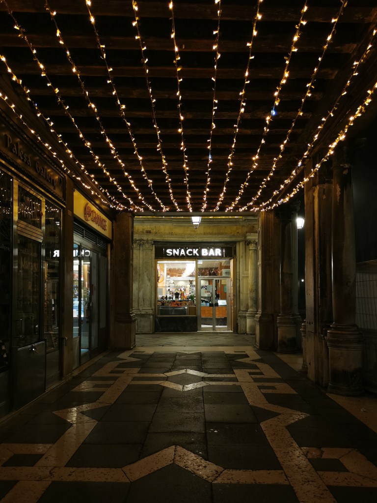 Around Piazza San Marco are covered alcoves with shops. We're looking at "SNACK BAR" with a simple cool yellow light sign and cool yellow lights inside. Above us are rows of warm yellow Christmas light decorations, leading us towards the shop.