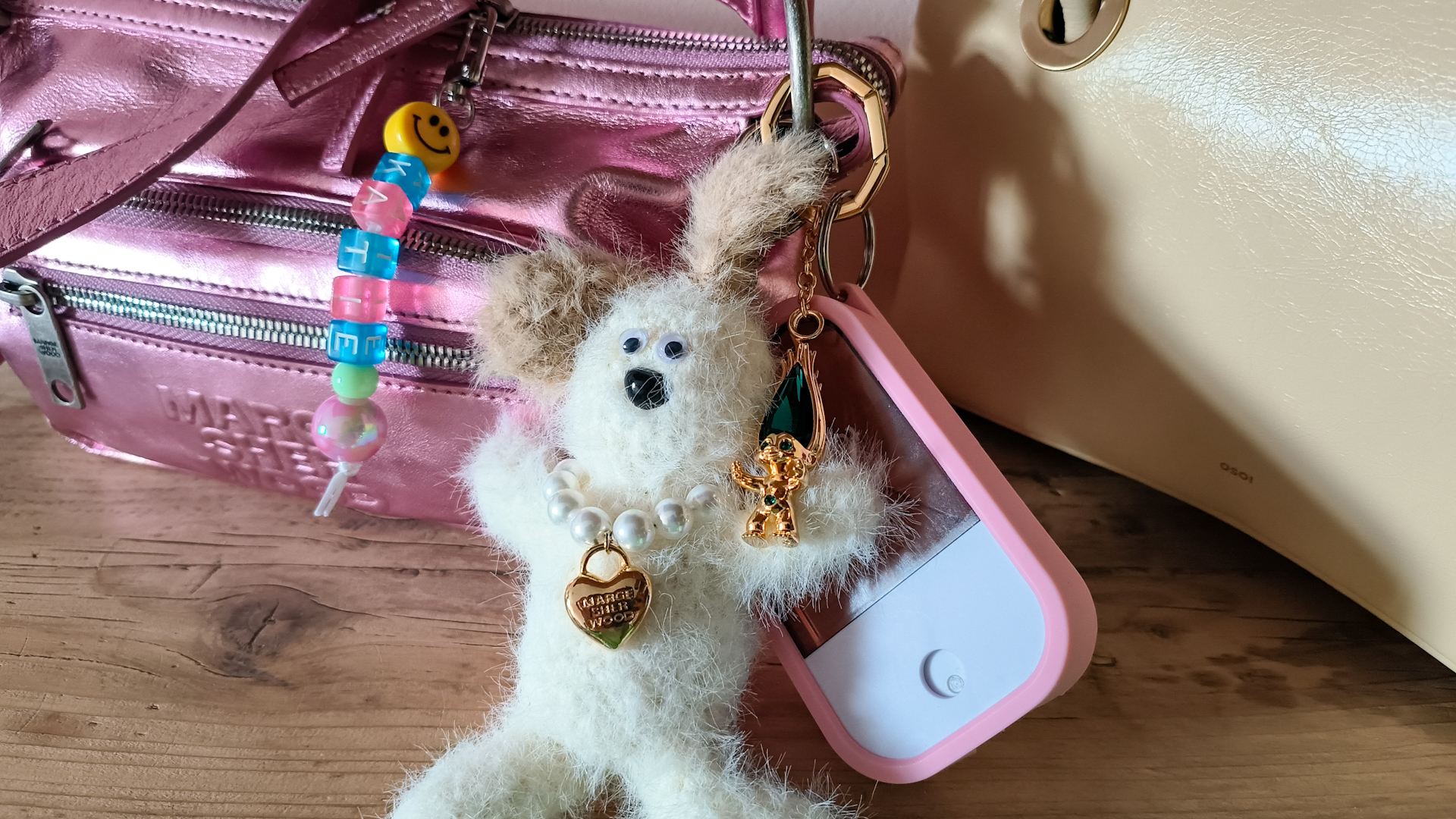 A small pink metallic handbag by Korean designer Marge Sherwood with personalised charms attached: a pink, blue and green beaded "Katie" name keyring made by an independent designer. A Marge Sherwood "Wood" keyring of a white dog with floppy brown ears, googly eyes and a pearl necklace with a gold heart charm. A Swarovski x DreamWorks Trolls keyring in gold tone with green crystals. And a Touchland hand sanitiser in a pink carrying case.