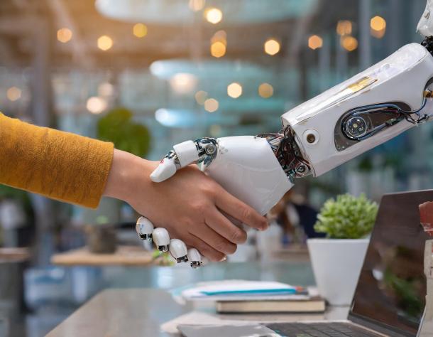 Image description, generated by Microsoft Copilot: A human hand and a robotic hand are shaking hands in a modern office setting. The human hand is wearing a yellow sleeve, while the robotic hand is white with visible mechanical components and wiring. In the background, there are blurred office elements such as a laptop, a plant, and some books on a desk. The image symbolizes the collaboration between humans and robots or artificial intelligence in a professional environment.